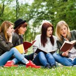 Four women reading books on a grassy field.