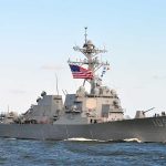 Military ship sails with American flag.