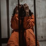 Person in orange jumpsuit sitting in a cell