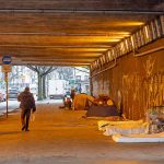 People walk under a bridge with homeless encampment.