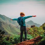 Child with backpack stands on mountain, arms outstretched.