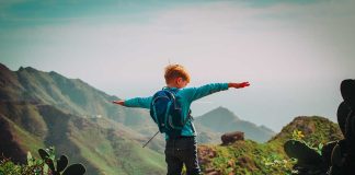 Child with backpack stands on mountain, arms outstretched.