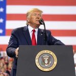 Man speaks at podium with U.S. flag background.