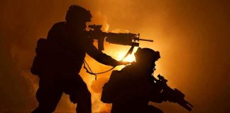 Two soldiers with weapons silhouetted against fiery background.