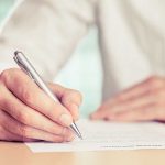A hand holding a pen writing on a document in an office setting