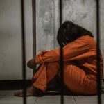 Person in orange jumpsuit sitting behind bars, head down.