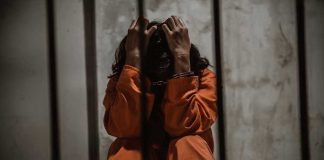 Person in orange jumpsuit sitting behind bars, handcuffed.