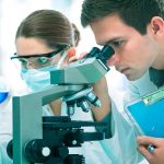 Two scientists conducting an experiment in a laboratory, one looking through a microscope and the other holding a flask with blue liquid