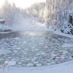 A serene winter landscape featuring a frozen lake surrounded by snow-covered trees and a log cabin