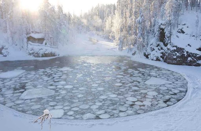 A serene winter landscape featuring a frozen lake surrounded by snow-covered trees and a log cabin