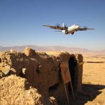 Military aircraft flying over a desert with an abandoned bunker and a for sale sign