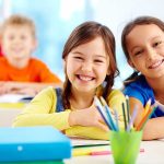 Two smiling girls in a classroom with colorful stationery and classmates in the background