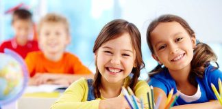 Two smiling girls in a classroom with colorful stationery and classmates in the background