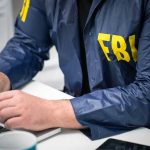 Person in FBI jacket typing on a laptop.
