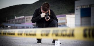 Crime scene photographer behind police tape with evidence marker.