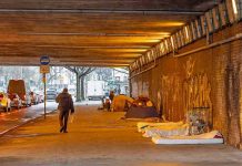 People walk under a bridge with homeless encampment.
