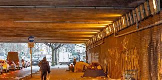 People walk under a bridge with homeless encampment.