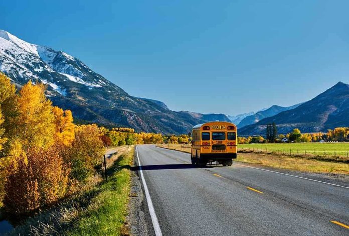 A yellow school bus driving on a rural road surrounded by autumn trees and mountains