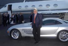 A man in a suit poses next to a silver luxury car in front of a private jet