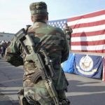 Blue-State Backlash Over D.C. Troops Soldier stands with rifle in front of American flag.