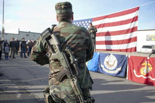 Soldier stands with rifle in front of American flag.