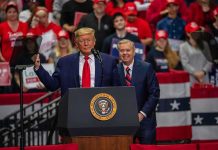 Trump’s Roast Turns Hostile: Press Left Shaken two men at a podium during a large indoor rally