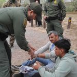 Illegal Caught by His Own Mugshot After Lying to ICE law enforcement officer handcuffing two seated men outdoors