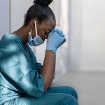 Healthcare worker in scrubs sitting against a wall with head bowed