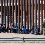 Group of people sitting by a tall border fence with an officer standing nearby